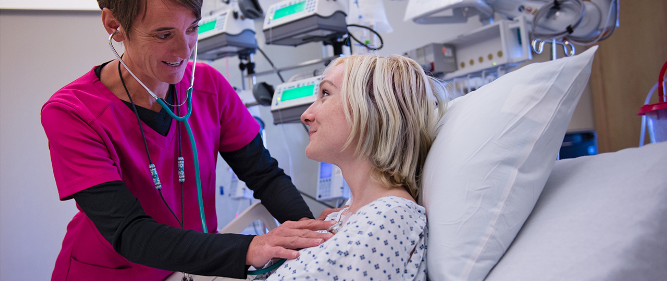 Nurse in pink scrubs checks the heartbeat of a patient. Both are smiling. Medical testing equipment is visible in the background.