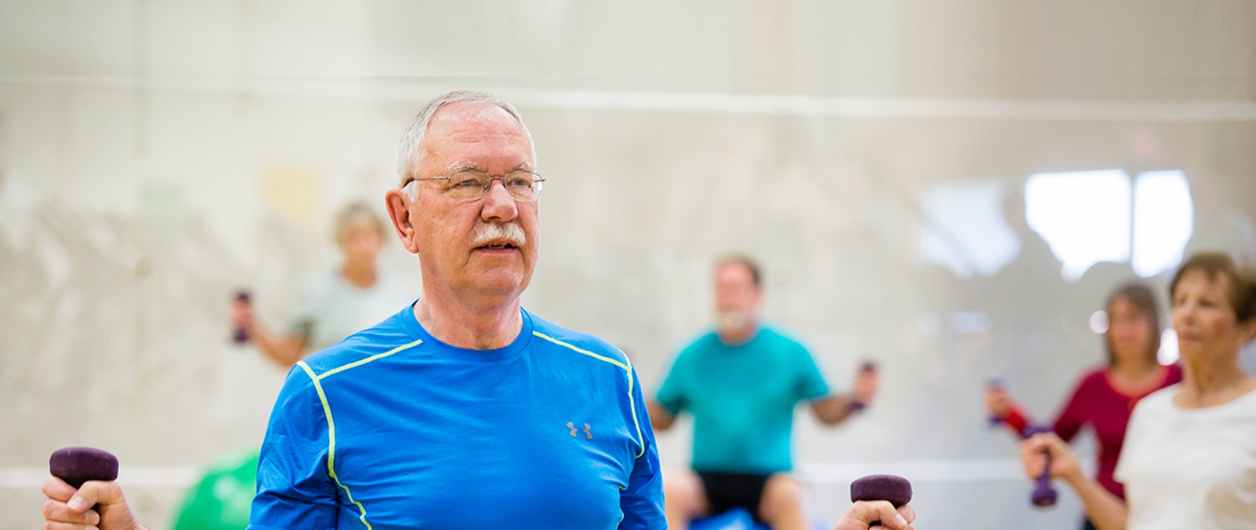 Exercise class with five people visible, standing with small dumbbells in each hand. The man in the foreground is older, with a gray mustache and glasses, blue workout shirt with green trim.