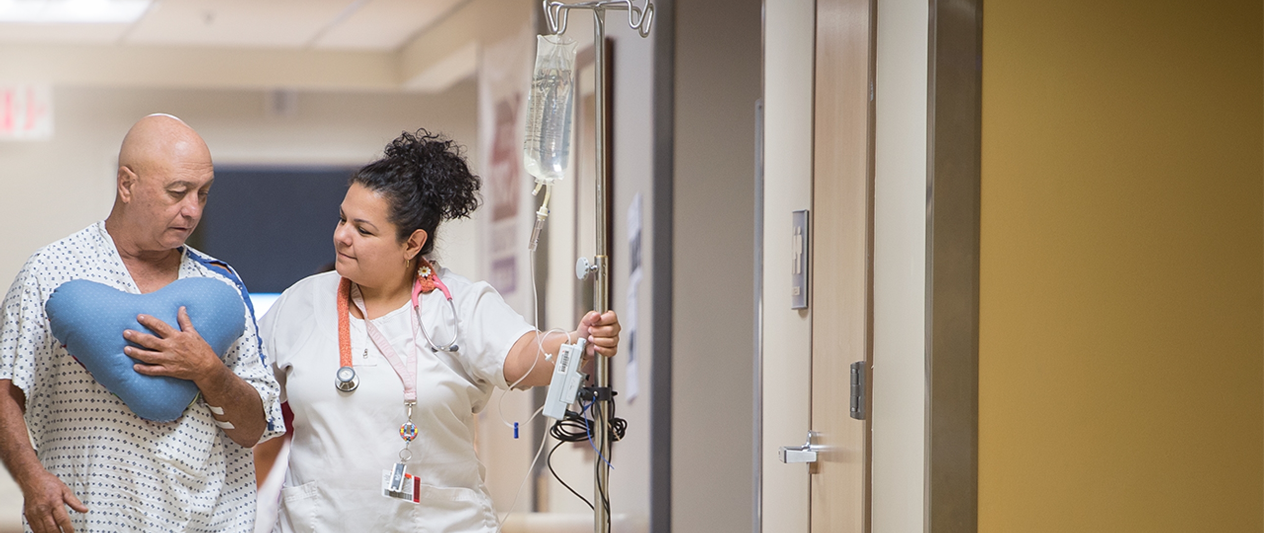 A nurse assists a male hospital patient walk down the hall. He clutches a heart-shaped pillow to his chest.