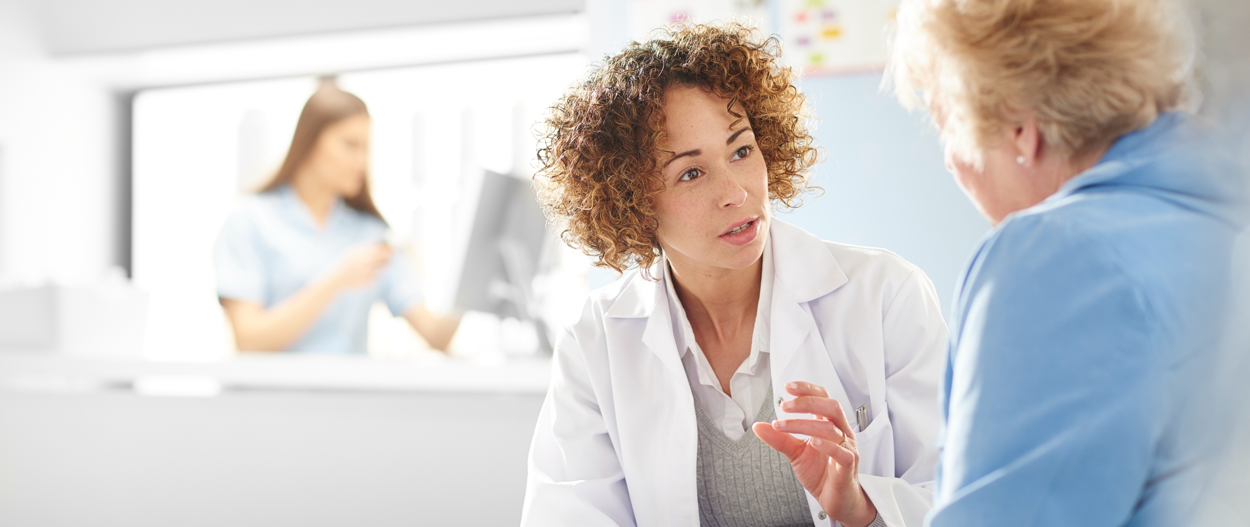 A doctor consults with  a patient in a hospital setting. In the background, an attendee stands behind a desk at a computer.
