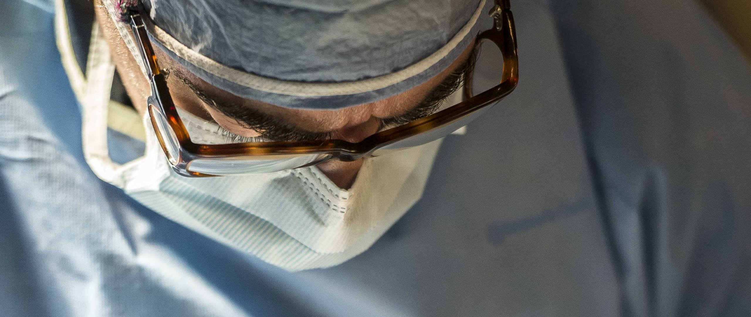 Close up of a male surgeon's face, wearing glasses and a mask, looking down. He appears to be intensely concentrating on his work.