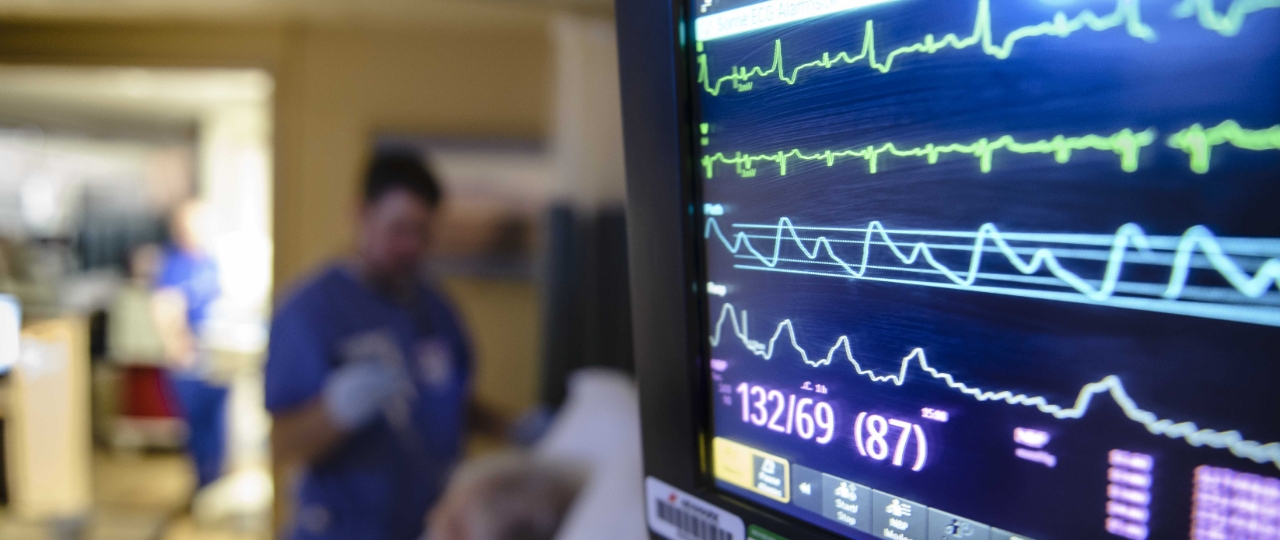 Hospital room, up close there is a screen monitoring several vital signs. Blood pressure reads 132/69. Out of focus, there is a nurse tending to the patient.