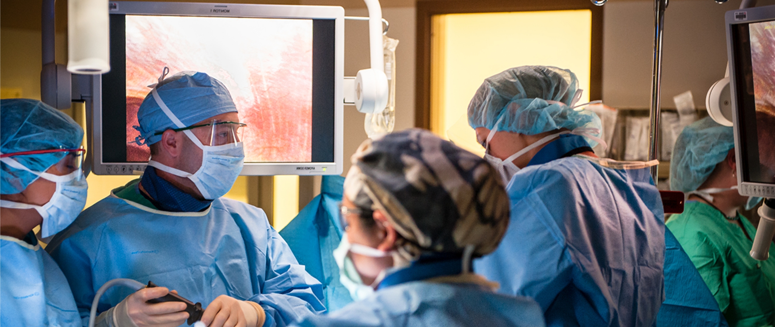Surgical setting, with five doctors and nurses in full scrubs, looking down. Patient is not visible, but two monitor screens showing the procedure are seen.